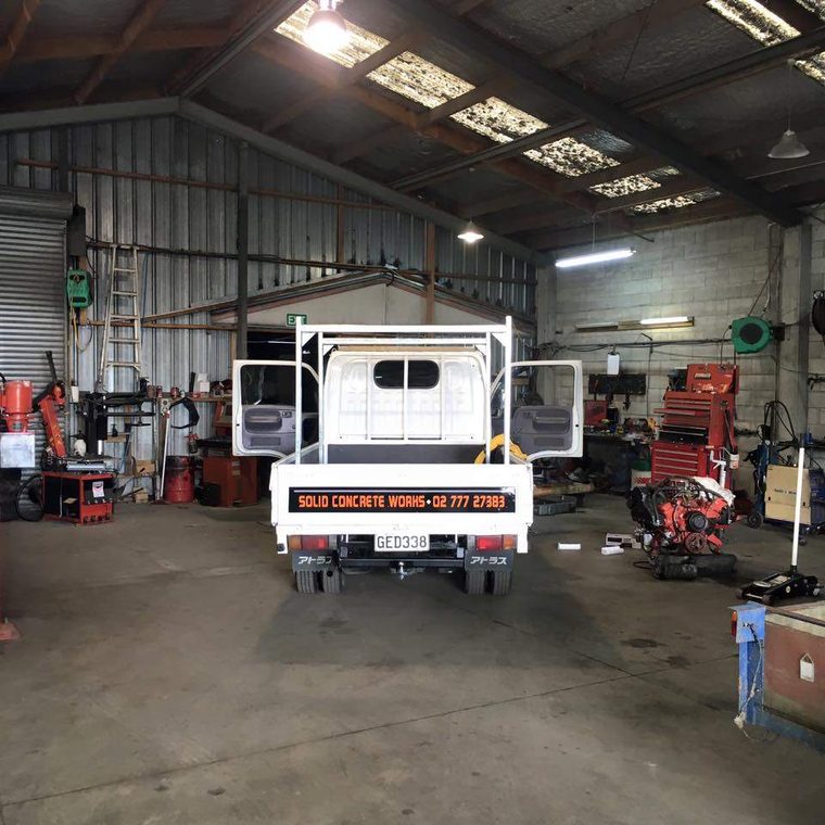 Rear view of a white light truck with its aluminium flat deck, parked in the middle of the workshop among tool chests and hoists.