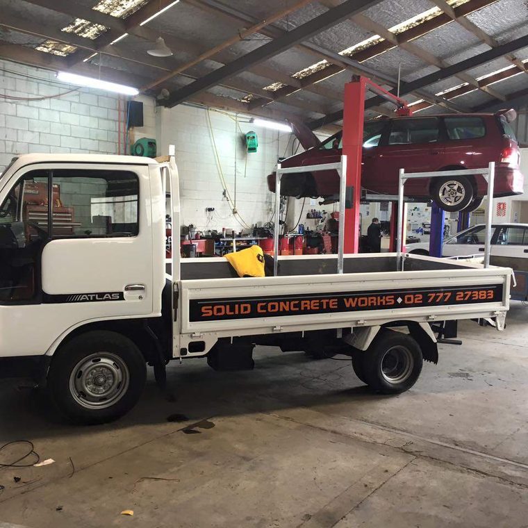A white light truck with an aluminium flat deck and a headboard rack, parked on the workshop floor under a red hoist.