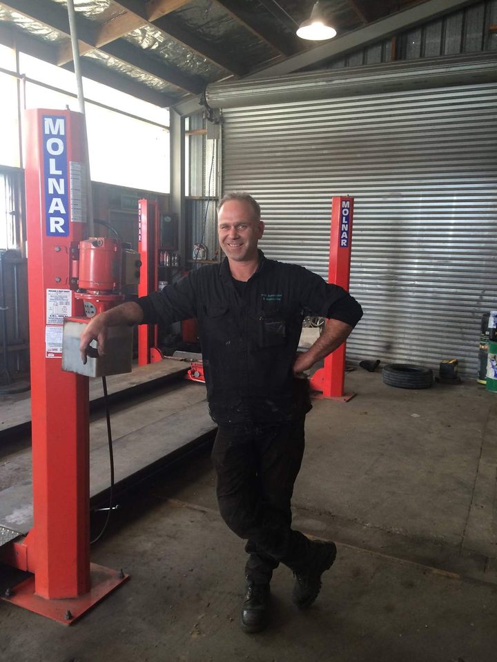 A mechanic in dark workwear standing beside a red two-post vehicle hoist inside the workshop, with the roller door behind him.