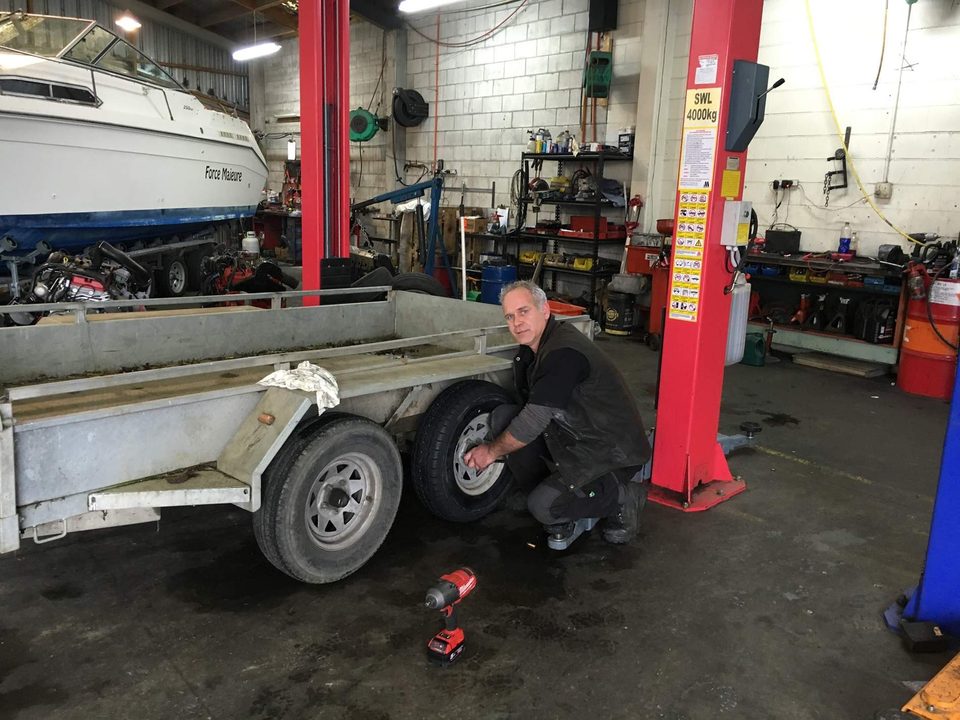 A mechanic crouched beside a galvanised tandem-axle trailer, fitting a wheel, with a boat and a red hoist behind him in the workshop.